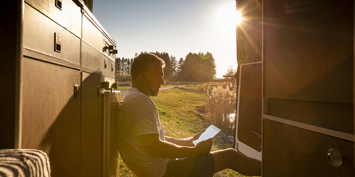 A man sitting in the open doorway of a van, reading a tablet in the warm sunlight with a scenic countryside in the background.