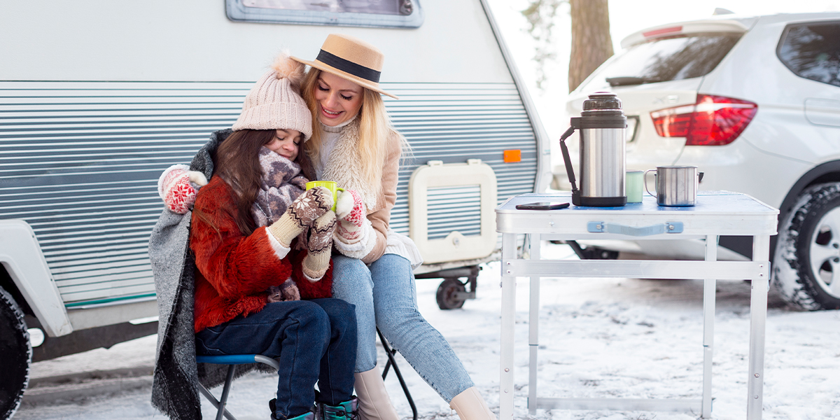 Mother and child enjoying hot drinks outside their RV at a snowy winter RV campground, dressed warmly in hats and scarves.
