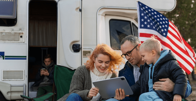 A family sitting outside their RV with an American flag, browsing on a tablet during Labor Day RV travel.