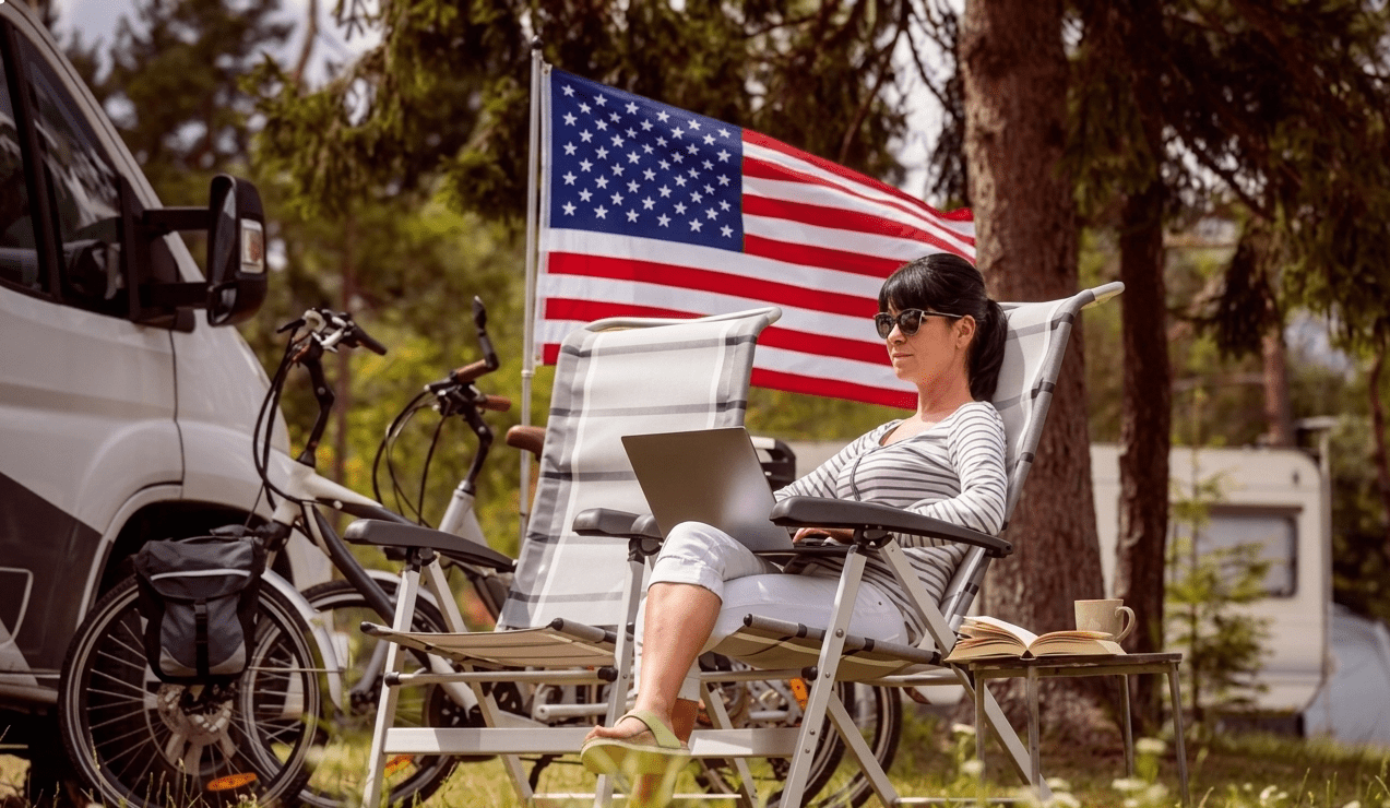 An RV traveler relaxing with a laptop at a campsite beside an American flag at the end of a Labor Day road trip.