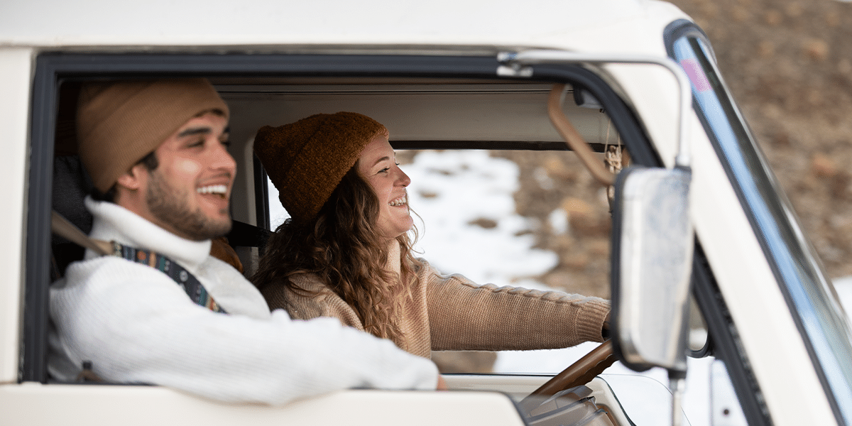 Smiling couple in winter hats driving an RV on a snowy road, heading south for snowbird season